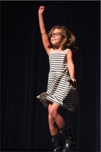 A little girl in a striped dress is jumping on stage.