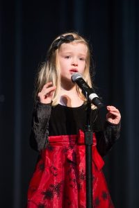 A little girl in a red dress singing into a microphone.