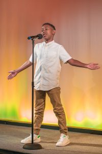 A boy is singing on stage with his arms outstretched.