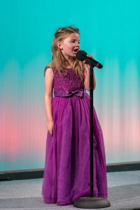 A little girl in a purple dress singing into a microphone.