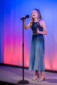 A girl in a blue dress singing into a microphone.