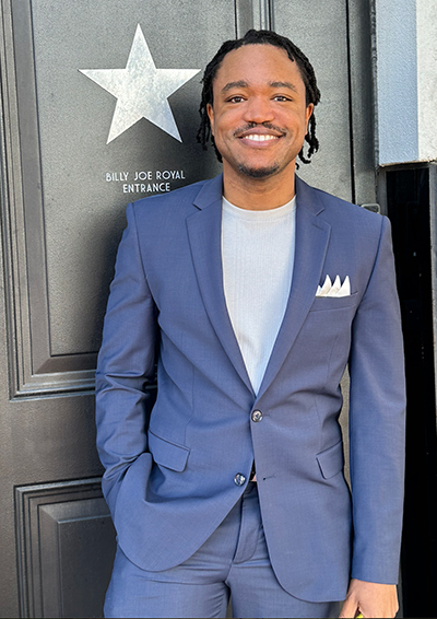 A man in a blue suit with a pocket square smiles next to the "billy joe royal entrance" sign with a star.