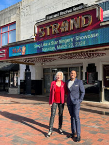 Two individuals posing in front of the strand theatre marquee advertising the "sing like a star singers' showcase" scheduled for saturday, march 23, 2024.