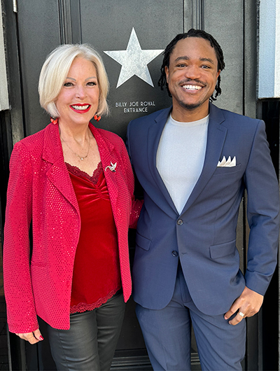 Two smiling individuals, one woman and one man, standing side by side in front of a door labeled "billy joe royal entrance" with a star symbol. the woman is wearing a red blazer and top, and the man is dressed in a blue suit with a white pocket square.