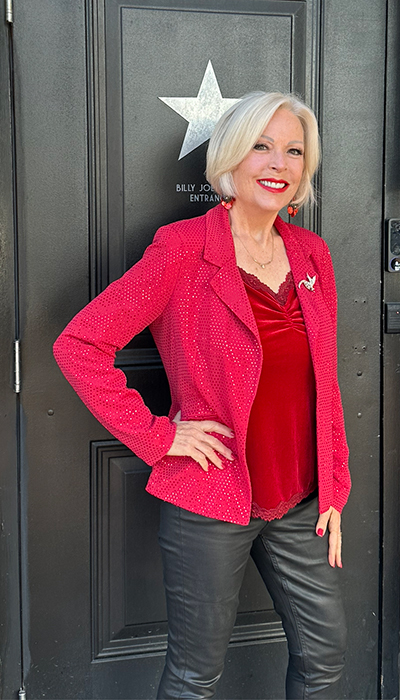 A woman standing by a door with a star sign, wearing a red sparkling jacket, a red top, and black trousers.