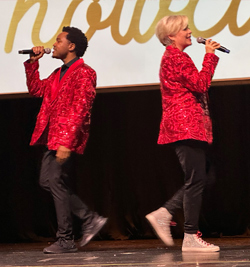 Two performers singing on stage, both wearing red patterned tops and black pants, with the male performer to the left and the female performer to the right.